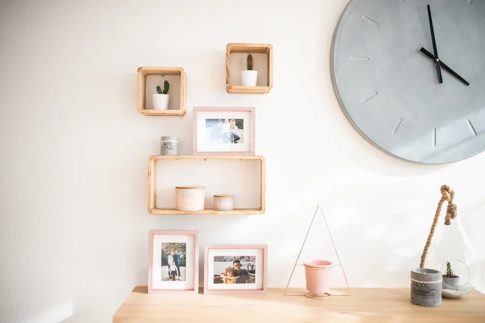 A bright white wall with small wooden shelves holding framed pictures and a plant, beside a round clock