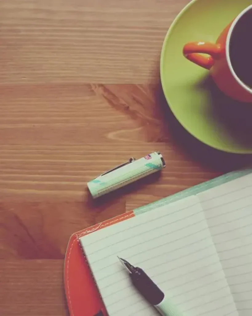 An open notebook with a pen beside a cup of tea on a wooden desk
