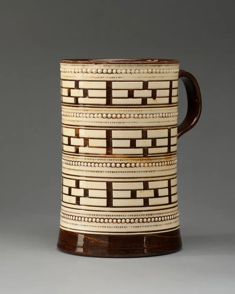 A cream ceramic mug with a brown geometric pattern and a dark rim, photographed on a light grey background
