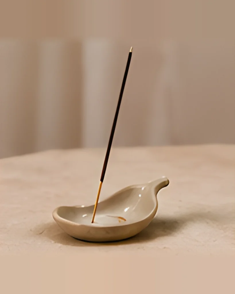 A small ceramic dish holding a stick of incense, with the incense burning