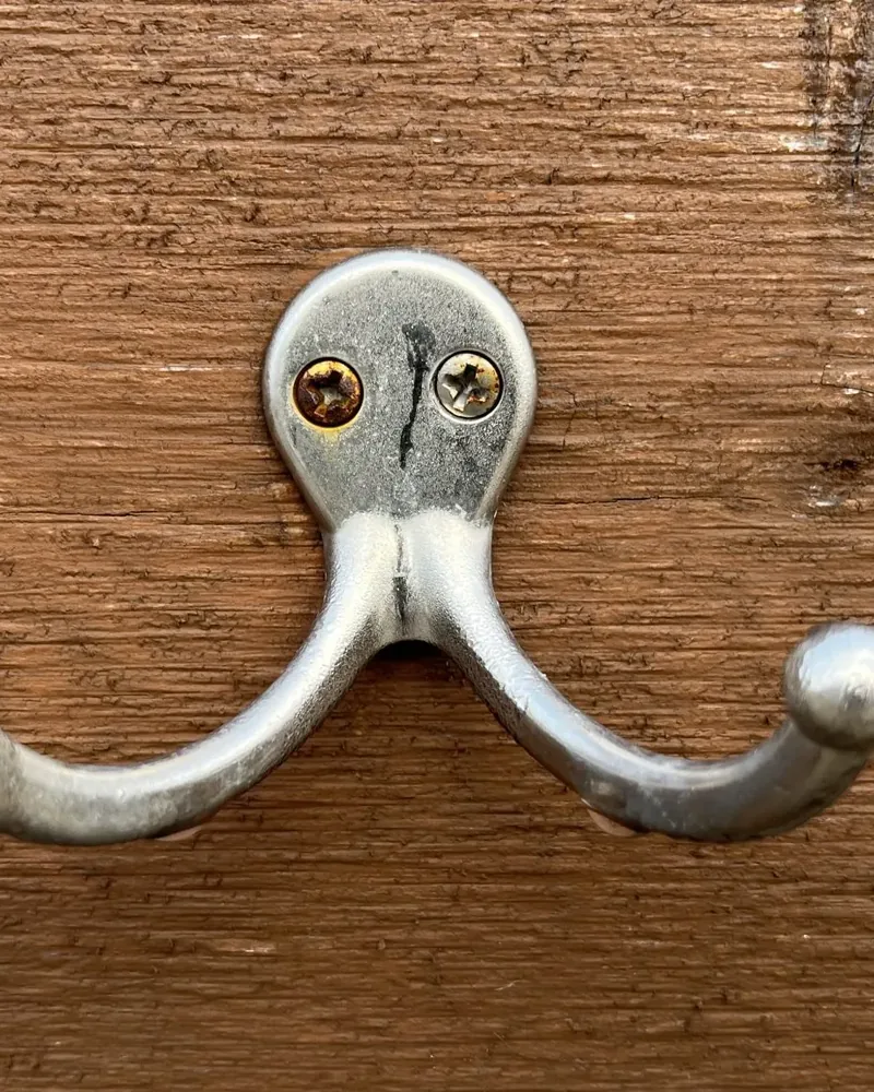 A metal double coat hook mounted on a wooden board