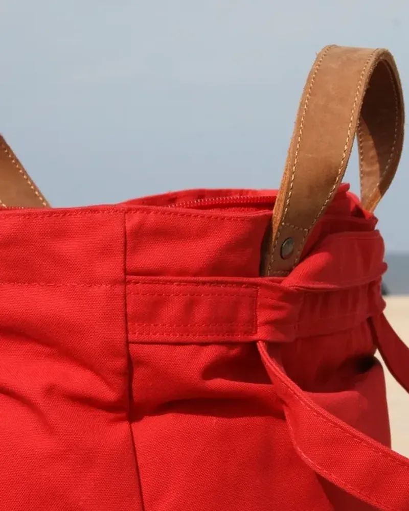 A red canvas tote bag with brown handles, photographed on a beach