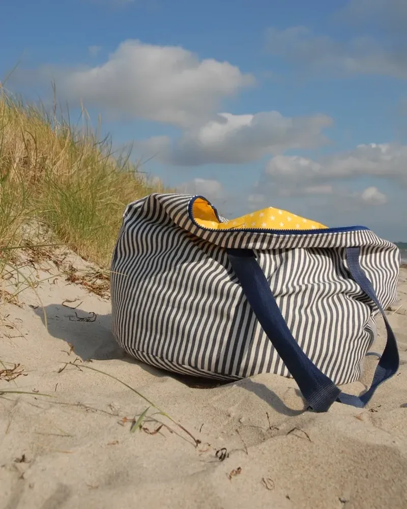 A striped canvas tote bag resting on beach sand under a blue sky