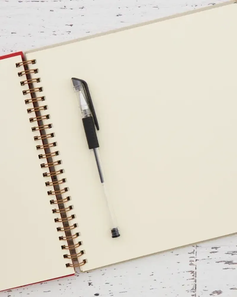 An open spiral-bound notebook with a pen and reading glasses on a white wooden desk